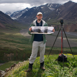 man standing on a hilltop near a camera, wide glacial valley in the distance
