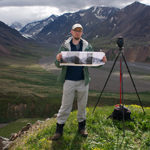 man standing on a hilltop near a camera, wide glacial valley in the distance man standing on a hilltop near a camera, wide glacial valley in the distance