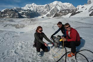 three people crouched near each other on a glacier under blue skies three people crouched near each other on a glacier under blue skies