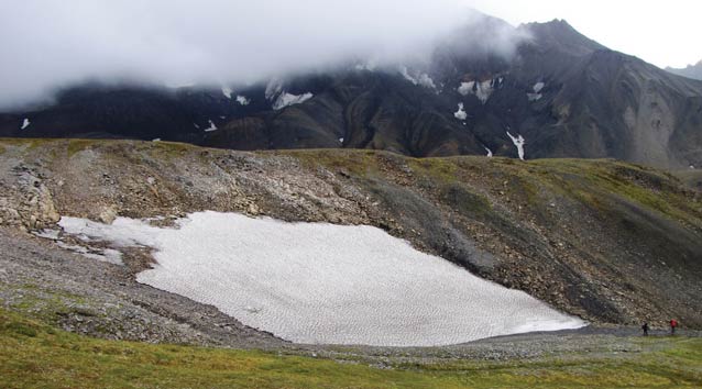 two people walking near a patch of snow on a mountain top two people walking near a patch of snow on a mountain top