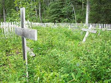 Wooden crosses stand at various angles in an overgrown plot, surrounded by a white picket fence. 