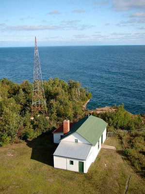 A radio antenna towers above a white, green-roofed building with blue water in the distance.