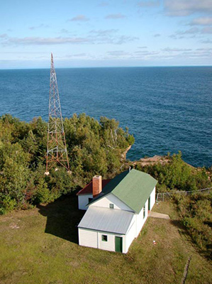 A radio antenna towers above a white, green-roofed building with blue water in the distance. A radio antenna towers above a white, green-roofed building with blue water in the distance.