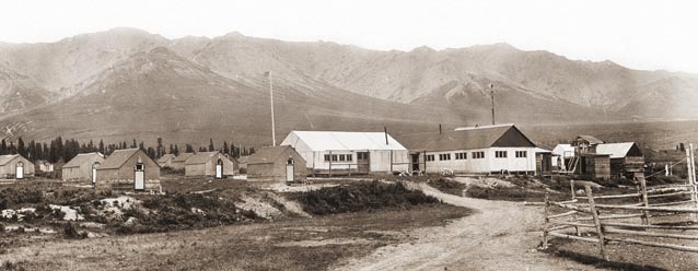 sepia toned image of numerous small cabins and two larger buildings with mountains in the distance