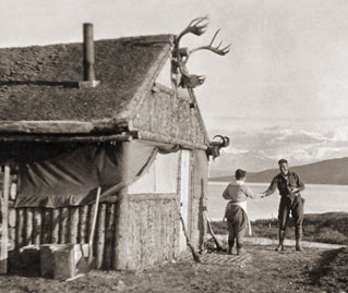 historic image of woman and man outside of a log cabin, with a lake and mountains in the distance