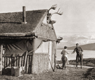 historic image of woman and man outside of a log cabin, with a lake and mountains in the distance historic image of woman and man outside of a log cabin, with a lake and mountains in the distance
