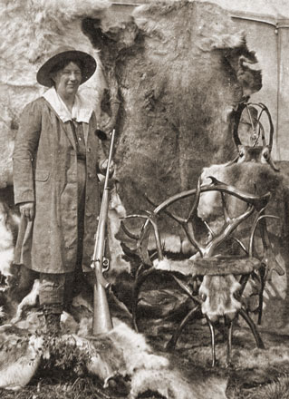 woman with a rifle posing next to animal skins and caribou antlers