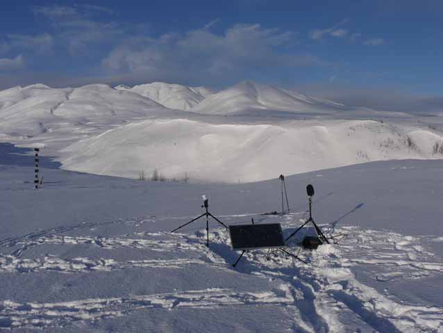 solar panel and microphones in a snowy fied, snow-covered hills in the distance