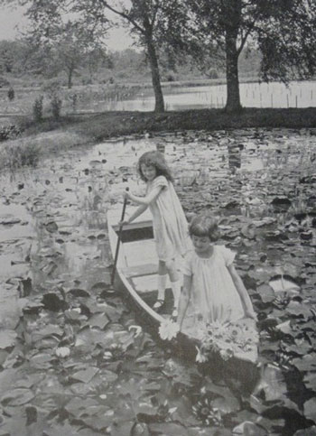 Two young girls in white dresses paddle through a pond dense with water lilies.