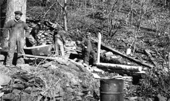 Men in work coveralls gather around the site of a dis-assembled building, built in a hillside.