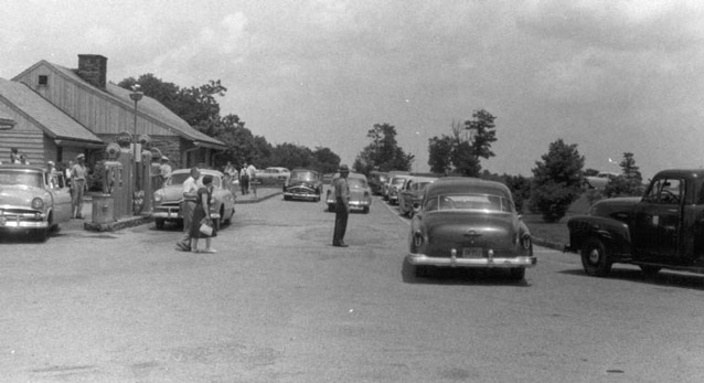 People and cars gather along the paved parking area in front of the service station building.