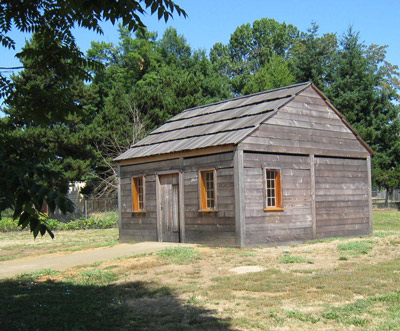 A small log house has a wood-shingled roof, several windows, and a door. 