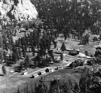 A high angle view of the landscape shows roads, structures and the row of cabins, and pine trees.