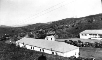 Large buildings, rows of smaller cabins, and trucks are arranged on the side of a slope.