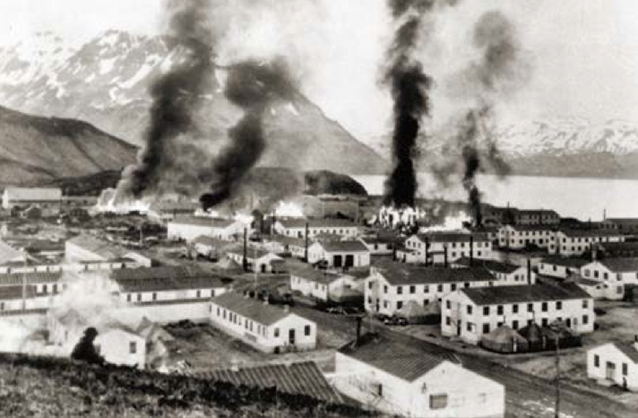View of buildings burning with four distinct plumes of black smoke.