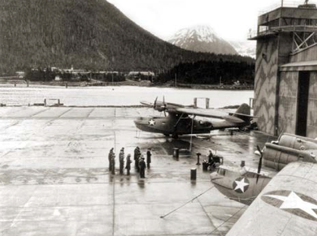 Military personnel stand on flight line in front of hangar near aircraft.