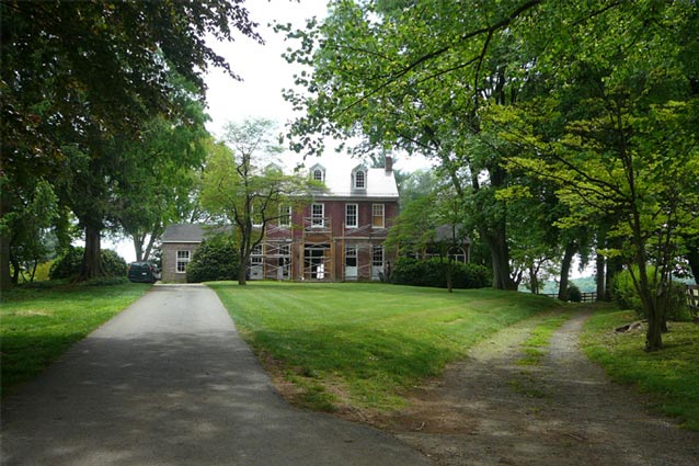 Gravel tracks curve to the right from a paved driveway, which leads towards a tall brick farmhouse.