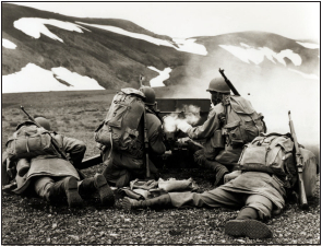 Four soldiers laying huddled behind a gun while firing.