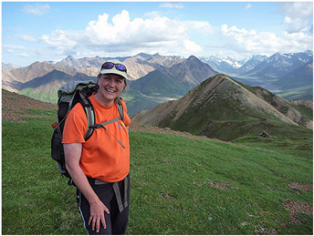 Woman wearing ballcap and daypack smiles while standing on green mountainscape