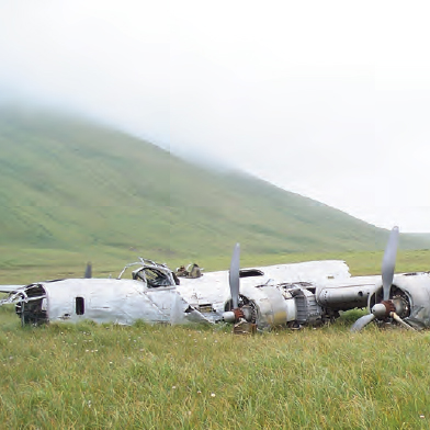 Color photo of plane wreckage in grass, hill beyond