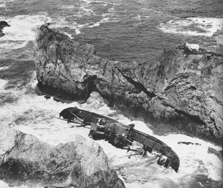 B&W photo of ship on its side amongst rocks and crashing waves