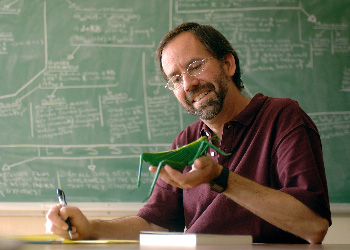 In front of a green chalkboard, a man makes handwritten notes from a scale model of a grasshopper