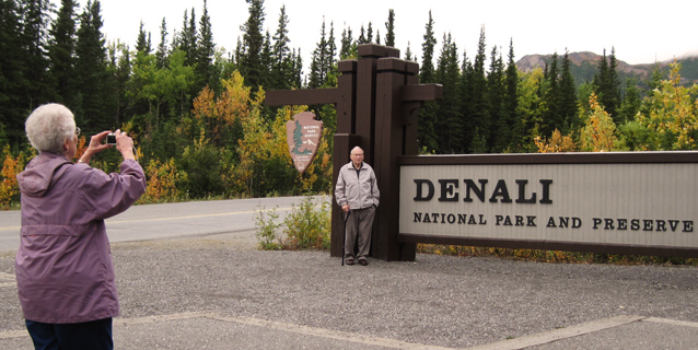a woman takes a picture of her husband standing by the Denali park entrance sign