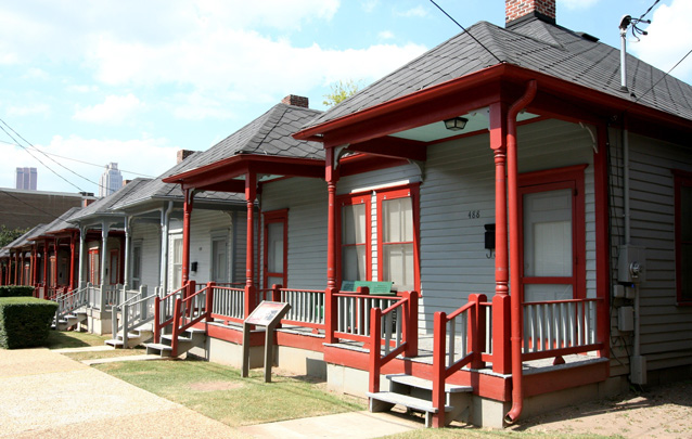 A row of single story houses with brightly painted front porch railings and small grass lots.