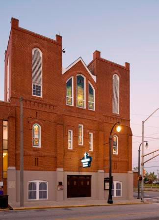 Light glows from the stained glass windows of a red brick church at twilight.