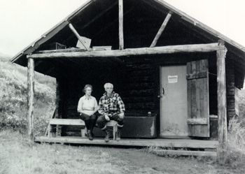Adolph and his wife sit in front on a log cabin Adolph and his wife sit in front on a log cabin