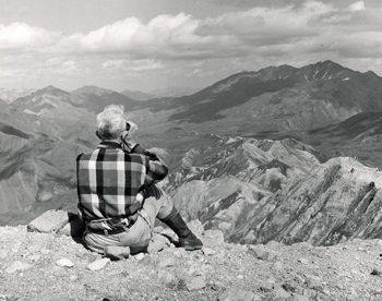 Murie sits on a rocky hillside looking out at the scenery while using binoculars