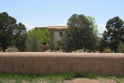 The adobe and stucco building is visible through trees, beyond a low adobe wall.