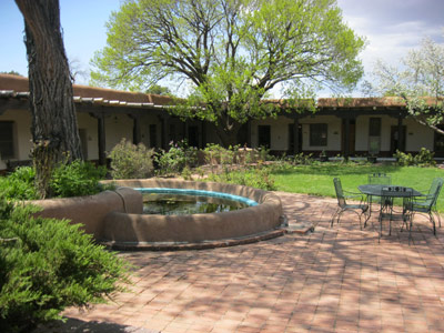 A built-in pond in a courtyard, surrounded by trees, an adobe and stucco building, and lawn.