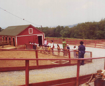 Several adults and a group of kids stand along a red fence by a barnyard. Several adults and a group of kids stand along a red fence by a barnyard.
