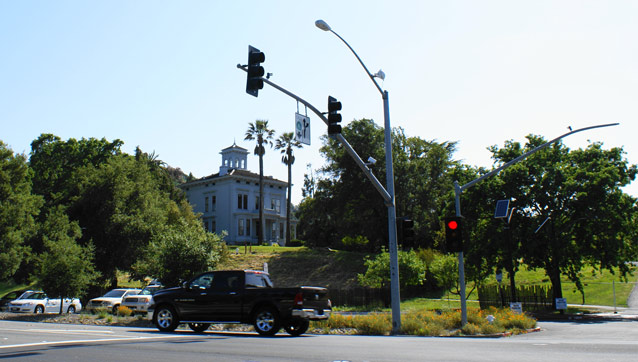 A large, rectangular house and leafy trees stand on a hill beyond traffic lights and a wide street.