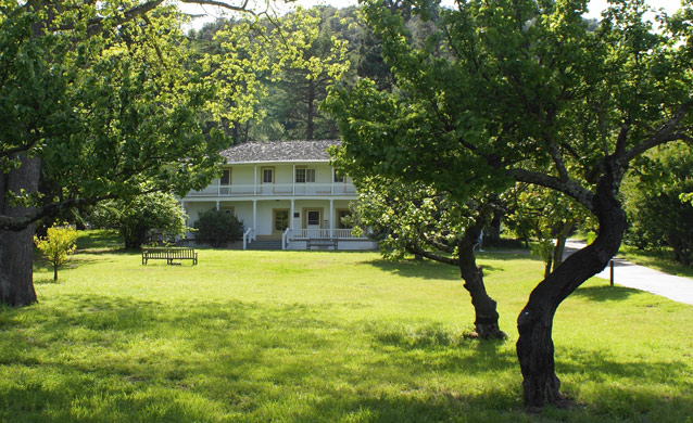 A two-story white house with a porch along both levels stands beyond orchards and lawn.