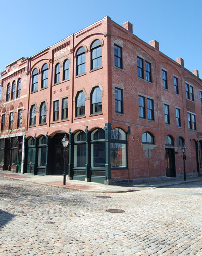 A three-story building of red brick and arching windows is beside a cobblestone street.