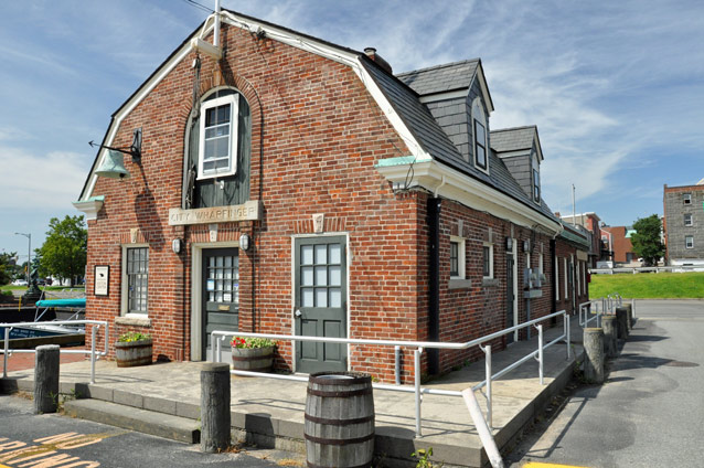 A red brick building has a rounded roof, two dormers, and a large bell hanging from the side.