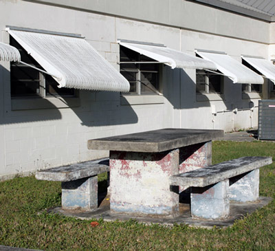 A solid concrete picnic table stands in grass next to a building with awnings over the windows.