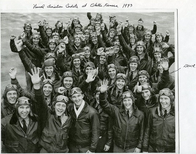 A large group of smiling men in aviator jackets, hats, and goggles raise their hands for the camera. A large group of smiling men in aviator jackets, hats, and goggles raise their hands for the camera.