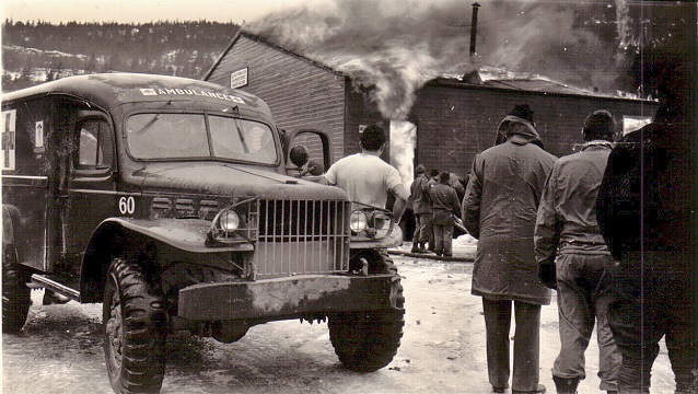 Men stand by ambulance watching a building burn.