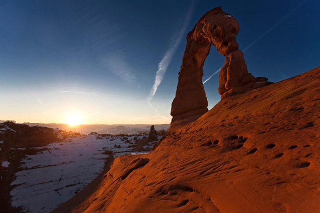 With snow covering the ground, the sun sits just below the horizon behind Delicate Arch.