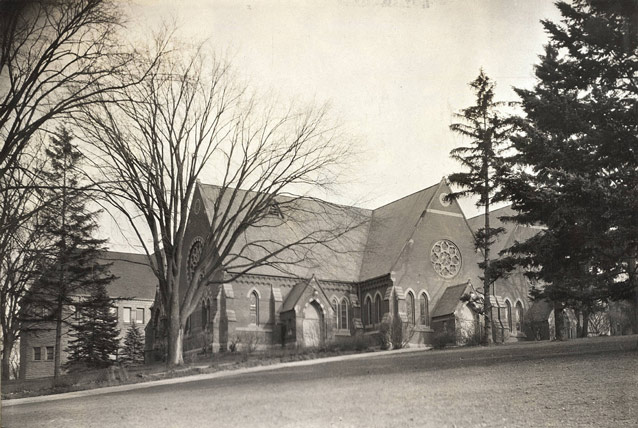 A chapel on a sloping lawn has a steeply pitched roof and round stained glass windows. 