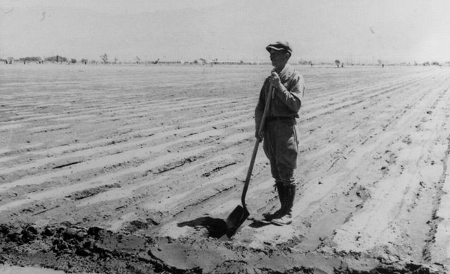 A man stands in the tip of his shovel resting in an irrigation ditch in a flat, open field.