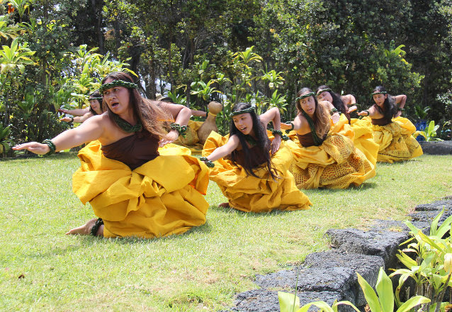 Women in traditional Hawai'ian dress crouch in a line to demonstrate a hula dance step