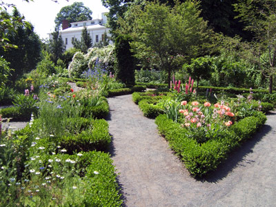 A gravel path leads around beds of colorful flowers, framed by boxwood hedges.