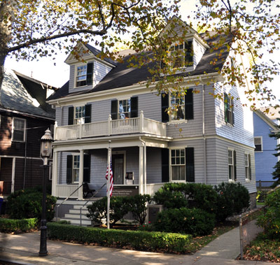 A two-story white house with a front porch on both levels, shrubs and sidewalk in front, and a flag.