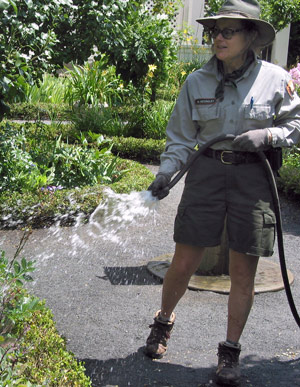 A woman in NPS uniform, in shorts and a hat, sprays water from a hose onto the edge of a garden.