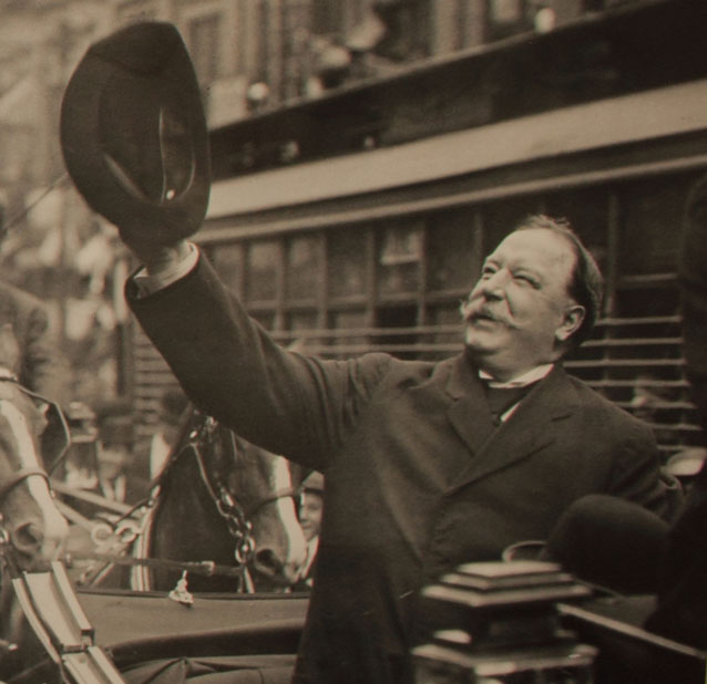 black and white photo of a man riding in an open car, waving his hat at a crowd