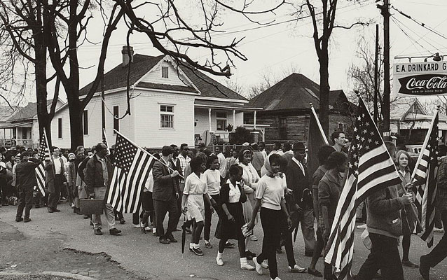 B&W; marchers with American flags walk in front of a white house.  Coca-cola sign hangs on right.